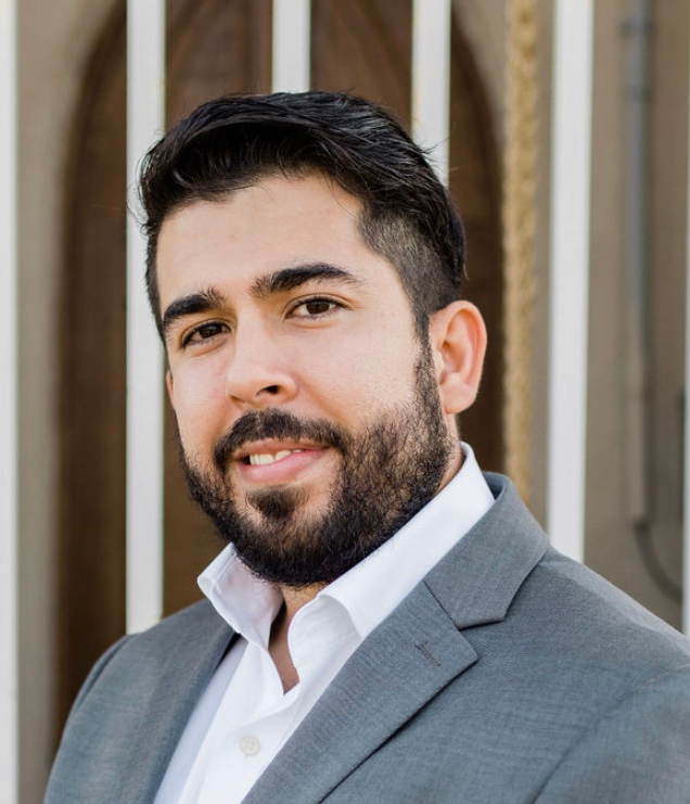 A person in a grey suit and white button down stands in front of white bars with a brown background.